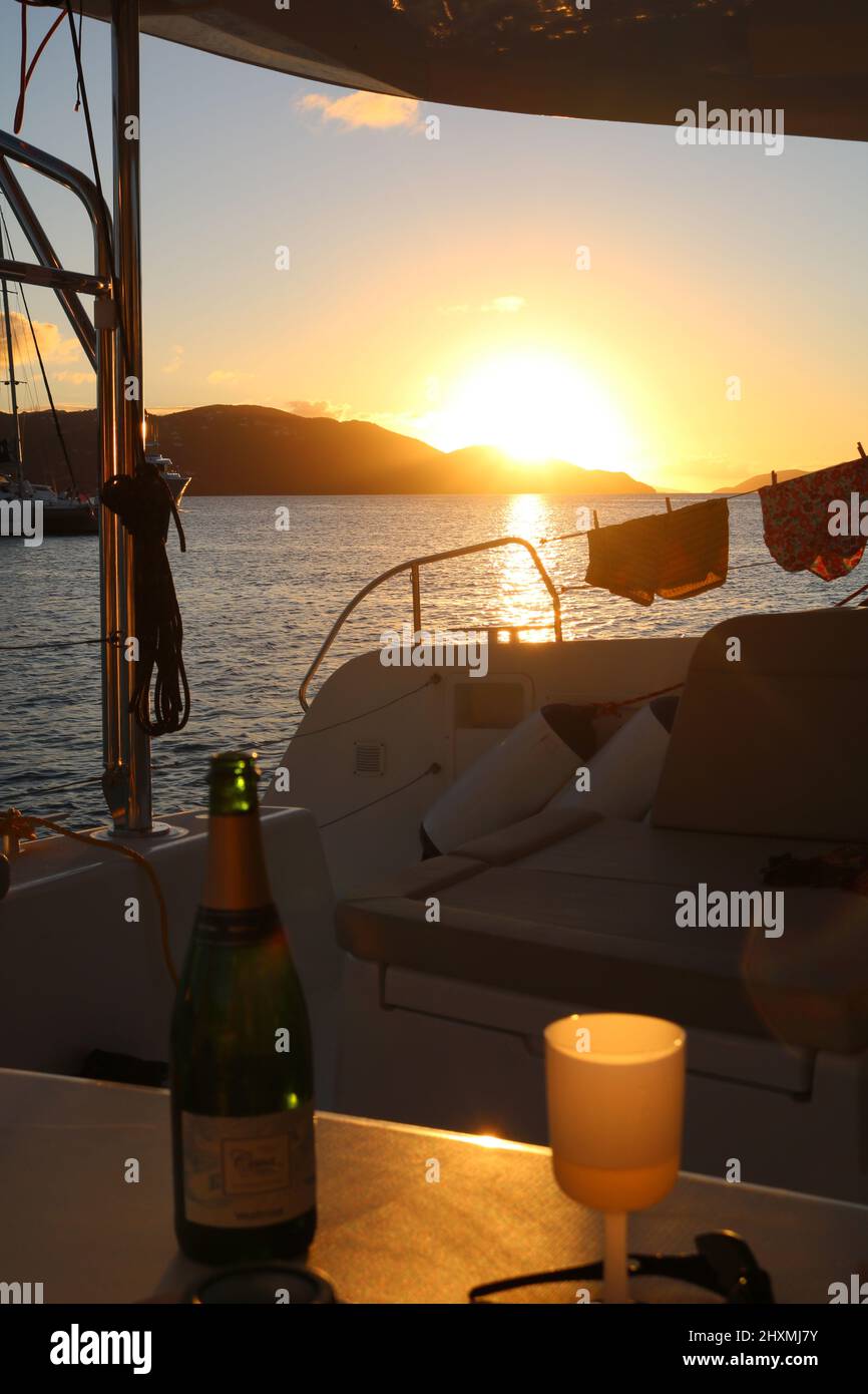 Champagner-Flasche und Glas, die gegen den Sonnenuntergang auf einer Yacht mit zwei Paar Badehosen auf den Sicherheitsdrähten der Yacht trocknen Stockfoto