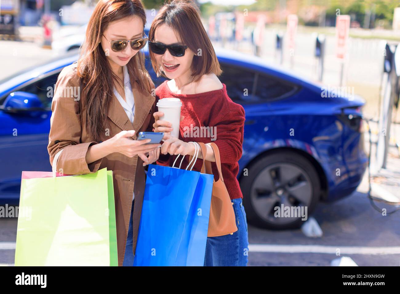 Glückliche junge Frau, die auf dem Stadtparkplatz in der Nähe eines Elektroautos steht, die Autobatterie von einer kleinen Stadtstation lädt, Einkaufstaschen hält und zuschaut Stockfoto