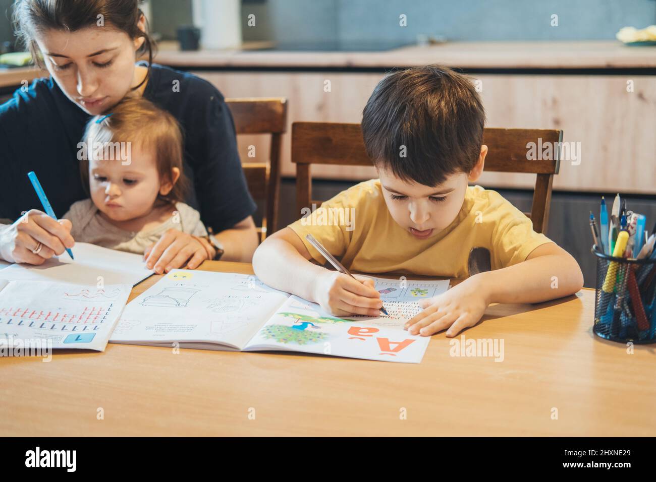 Junge, der am Tisch sitzt und das Schreiben praktiziert, Mutter hält das Schreiben eines kleinen Mädchens auf einem Laken konzentriert. Kindererziehung. Studieren, Hausaufgaben machen. Stockfoto