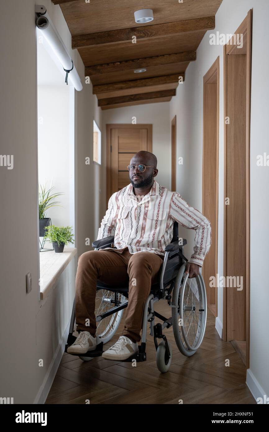 Afroamerikanischer Mann mit Behinderung sitzt auf dem Rollstuhl in einem langen Korridor einer großen modernen Wohnung und schaut auf die Kamera Stockfoto