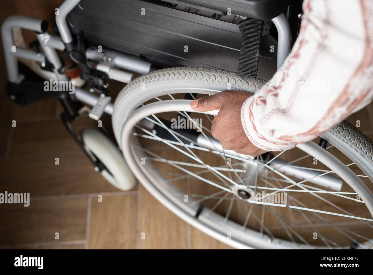 Blick über die Hand eines jungen Mannes mit Behinderung, der das Rad des Rollstuhls dreht, während er sich auf dem Holzboden hinunterbewegt Stockfoto