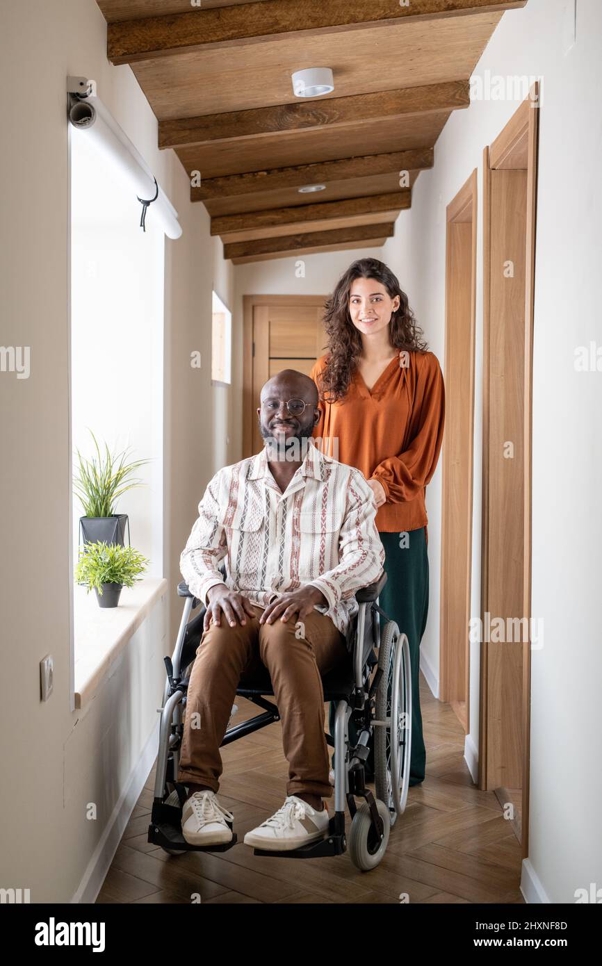 Junger, fröhlicher Mann mit Behinderung und seine Pflegekraft in der Freizeitkleidung, der im Korridor der modernen Wohnung die Kamera ansah Stockfoto