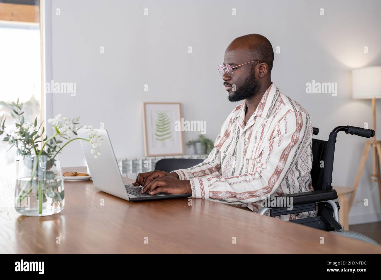 Junger seriöser Geschäftsmann oder Freiberufler im Rollstuhl sitzt am Tisch im Wohnzimmer und schaut beim Tippen auf den Laptop-Bildschirm Stockfoto