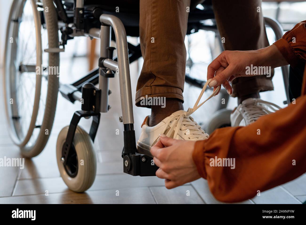 Hände einer jungen Betreuerin, die Schuhwerk auf die Schuhe eines Mannes mit Behinderung bindet, der auf dem Rollstuhl sitzt, bevor er zum Gehen geht Stockfoto