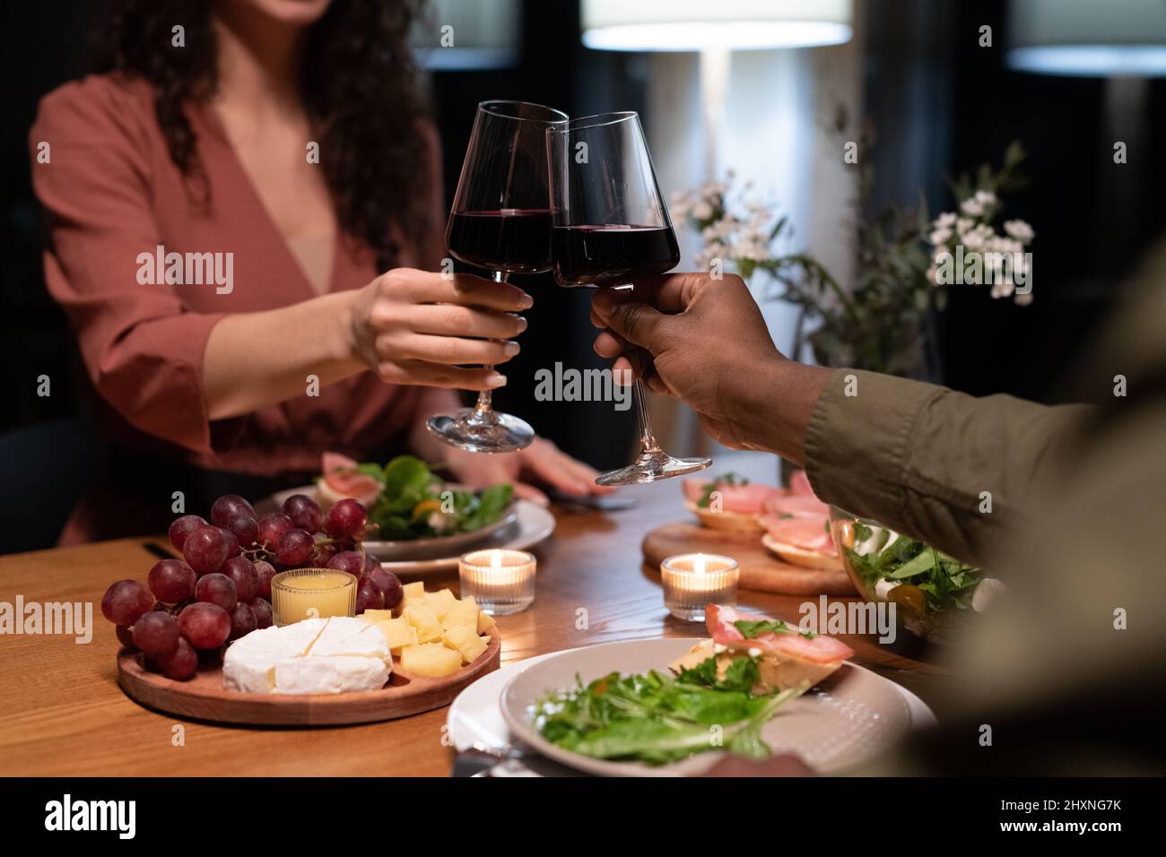 Die Hände junger gemischter Datteln, die mit Gläsern Rotwein über dem Esstisch klirren, werden mit gesunden Speisen und Snacks serviert Stockfoto