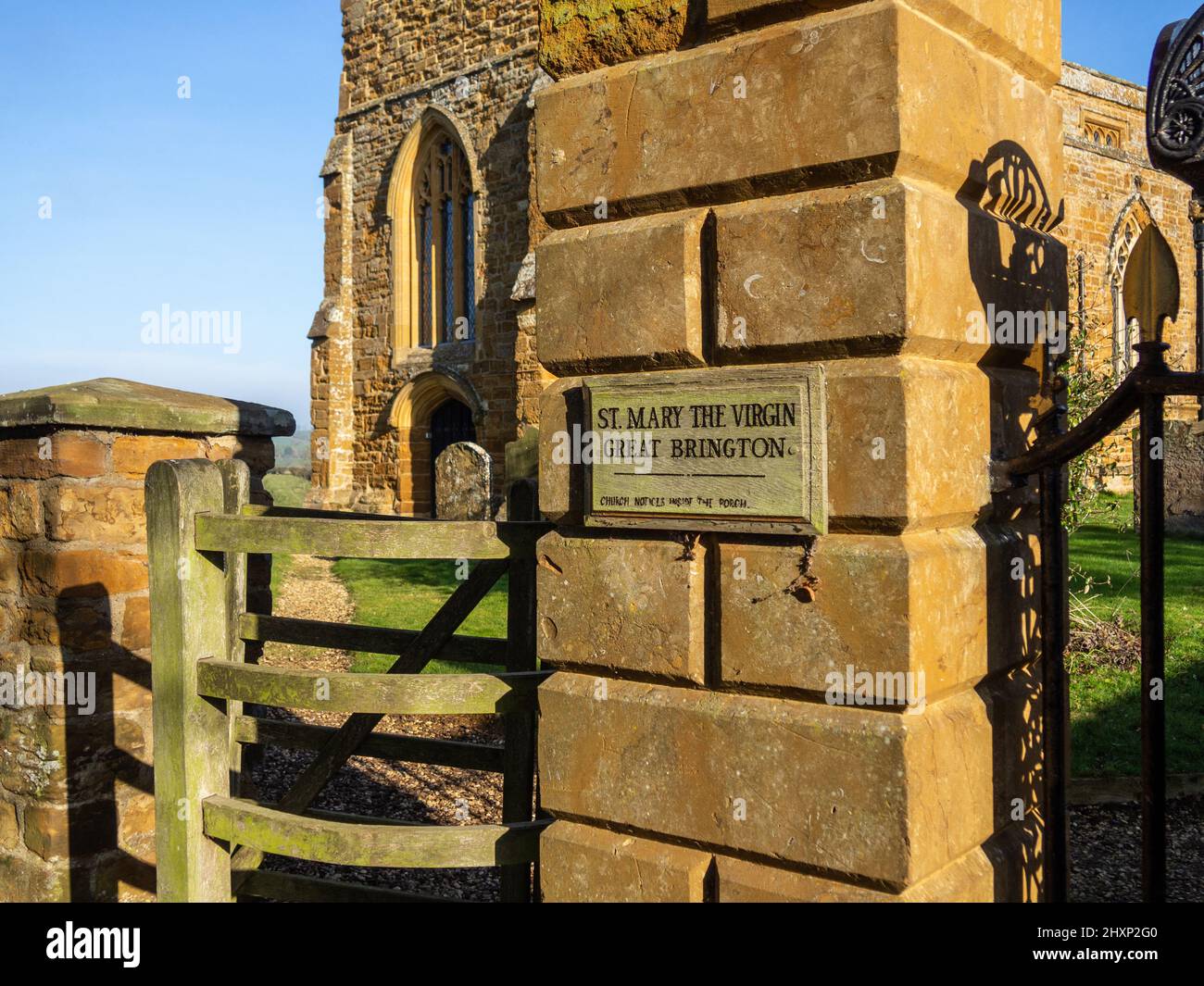 Holzschild für die Kirche St. Mary the Virgin, Great Brington, Northamptonshire, UK; befestigt auf einem Steinpier mit Kirche im Hintergrund. Stockfoto