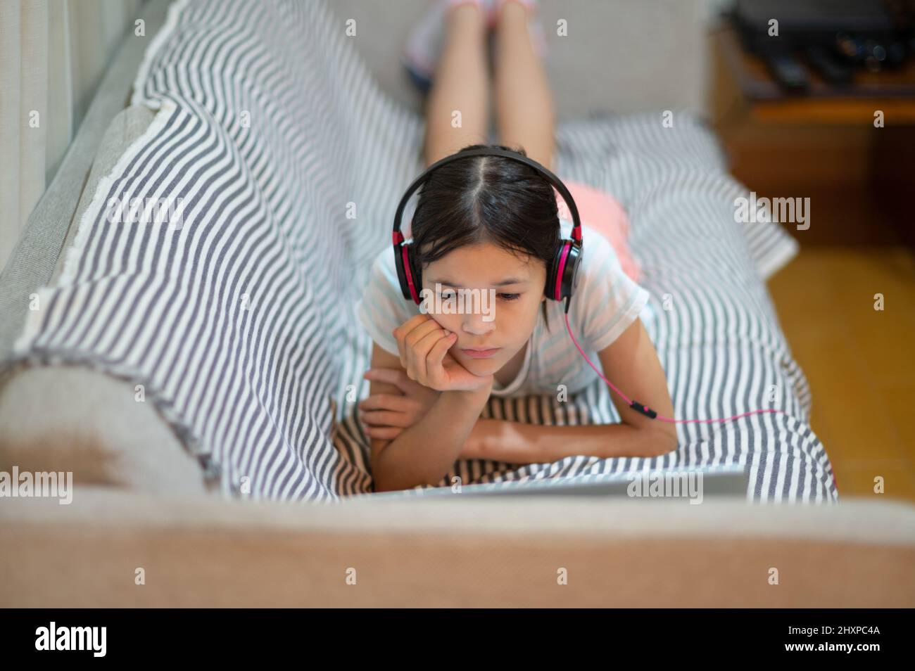 Junges eurasisches Mädchen, das auf dem Sofa liegt, Musik über Kopfhörer hört und einen Laptop benutzt. Stockfoto