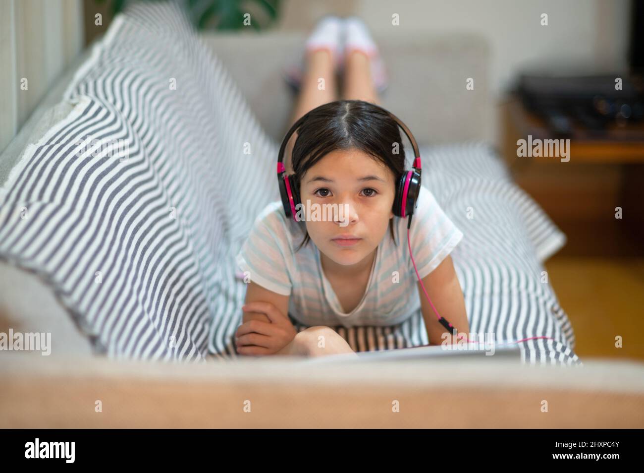 Süßes eurasisches junges Mädchen, Kind, das auf dem Sofa liegt und Musik über Kopfhörer hört und einen Laptop benutzt. Stockfoto