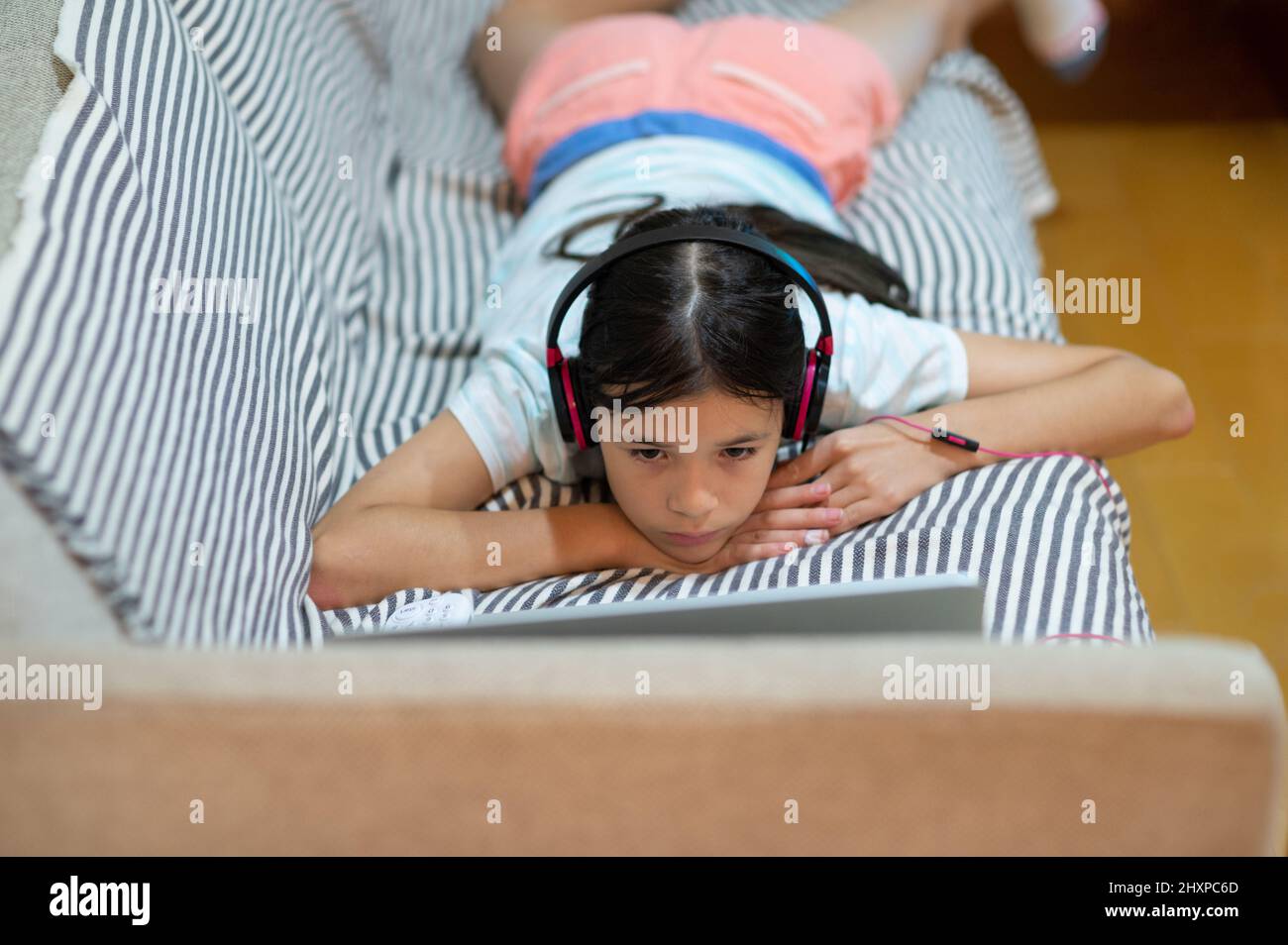 Junges eurasisches Mädchen, Kind, das auf dem Sofa liegt und Musik über Kopfhörer hört und einen Laptop benutzt. Stockfoto