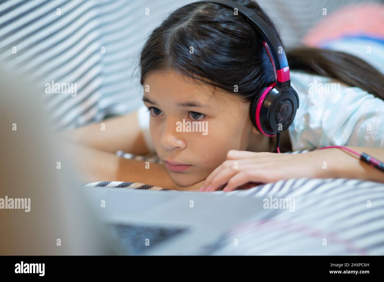 Nahaufnahme eines Mädchens, eines Kindes, das auf dem Sofa liegt und Musik über Kopfhörer hört und einen Laptop benutzt. Stockfoto