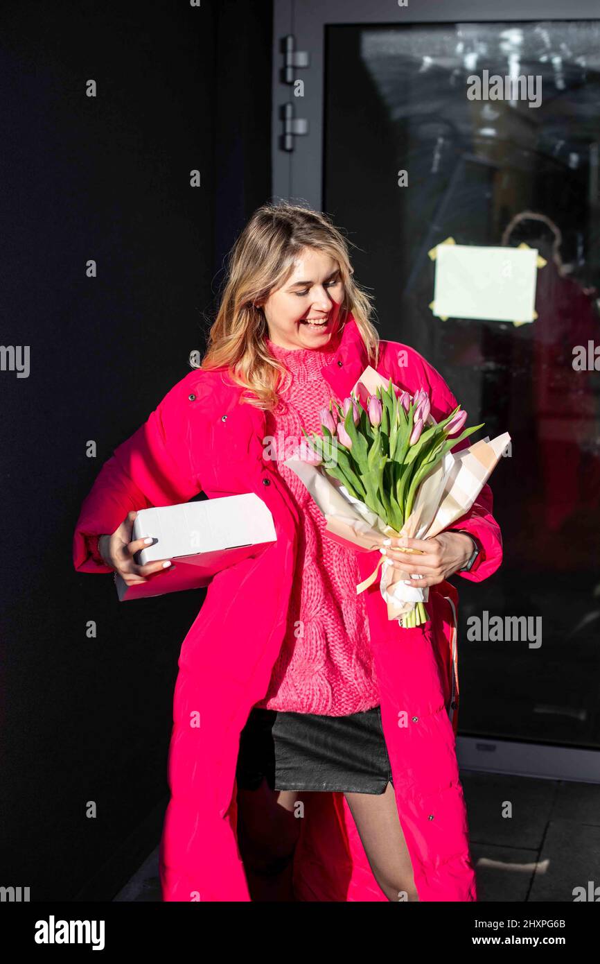 Vertikales Porträt der glücklichen lachenden blonden Frau in rosa Outfit, Geschenke, Blumenstrauß Tulpen. Firmenfeier zu Fuß. Stockfoto