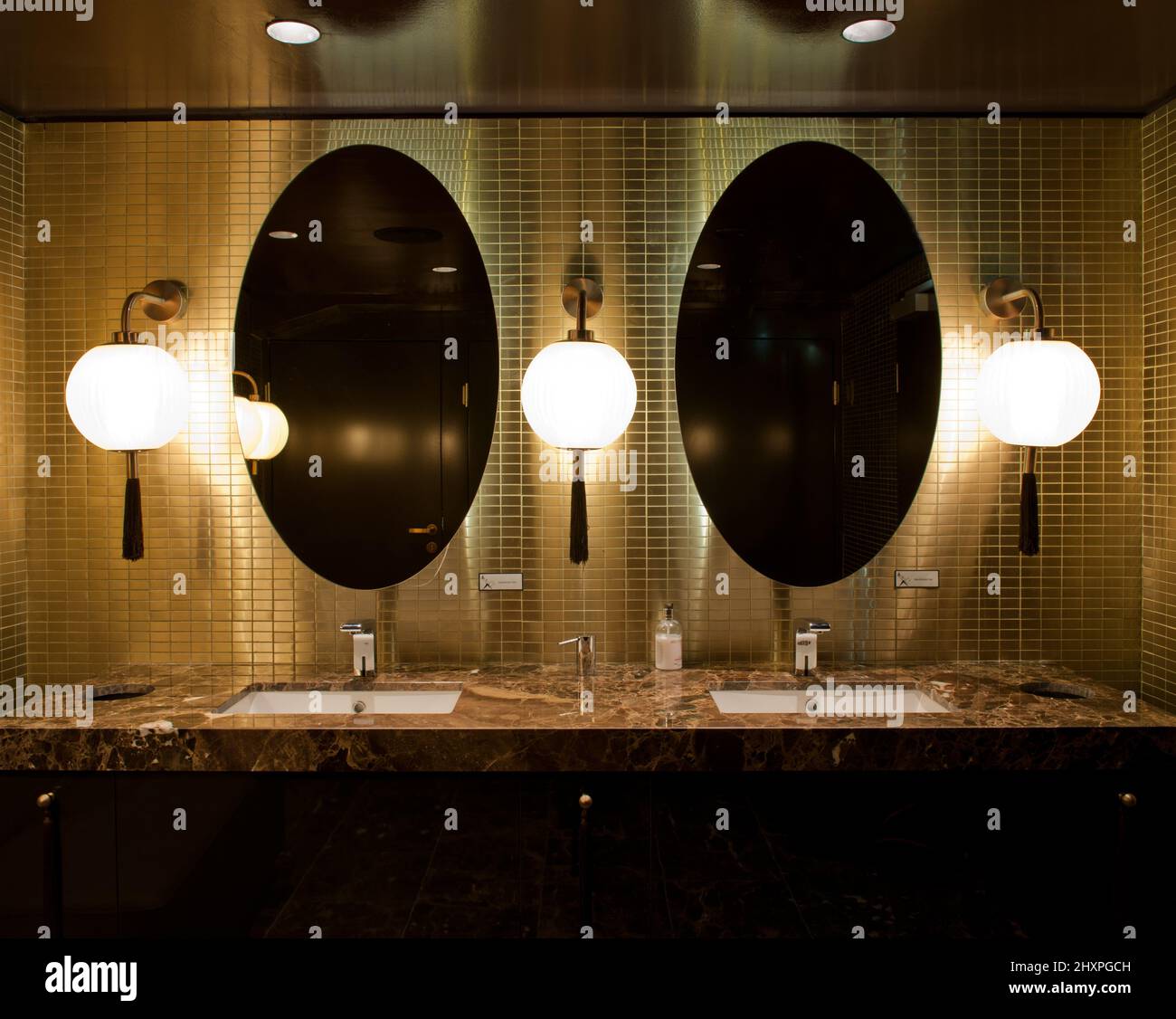 Zwei ovale Spiegel in goldgefliester Wand mit Waschbecken aus braunem Marmor und drei chinesischen Lampen Stockfoto