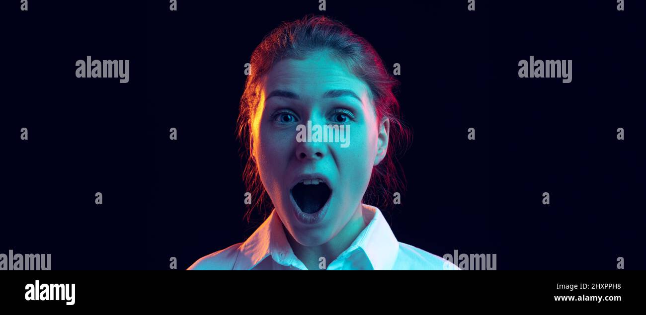 Schockiert junges Mädchen in weißem Hemd Blick auf die Kamera mit offenem Mund isoliert auf dunklem Hintergrund in blauem Neonlicht, Filter. Konzept der Emotionen Stockfoto