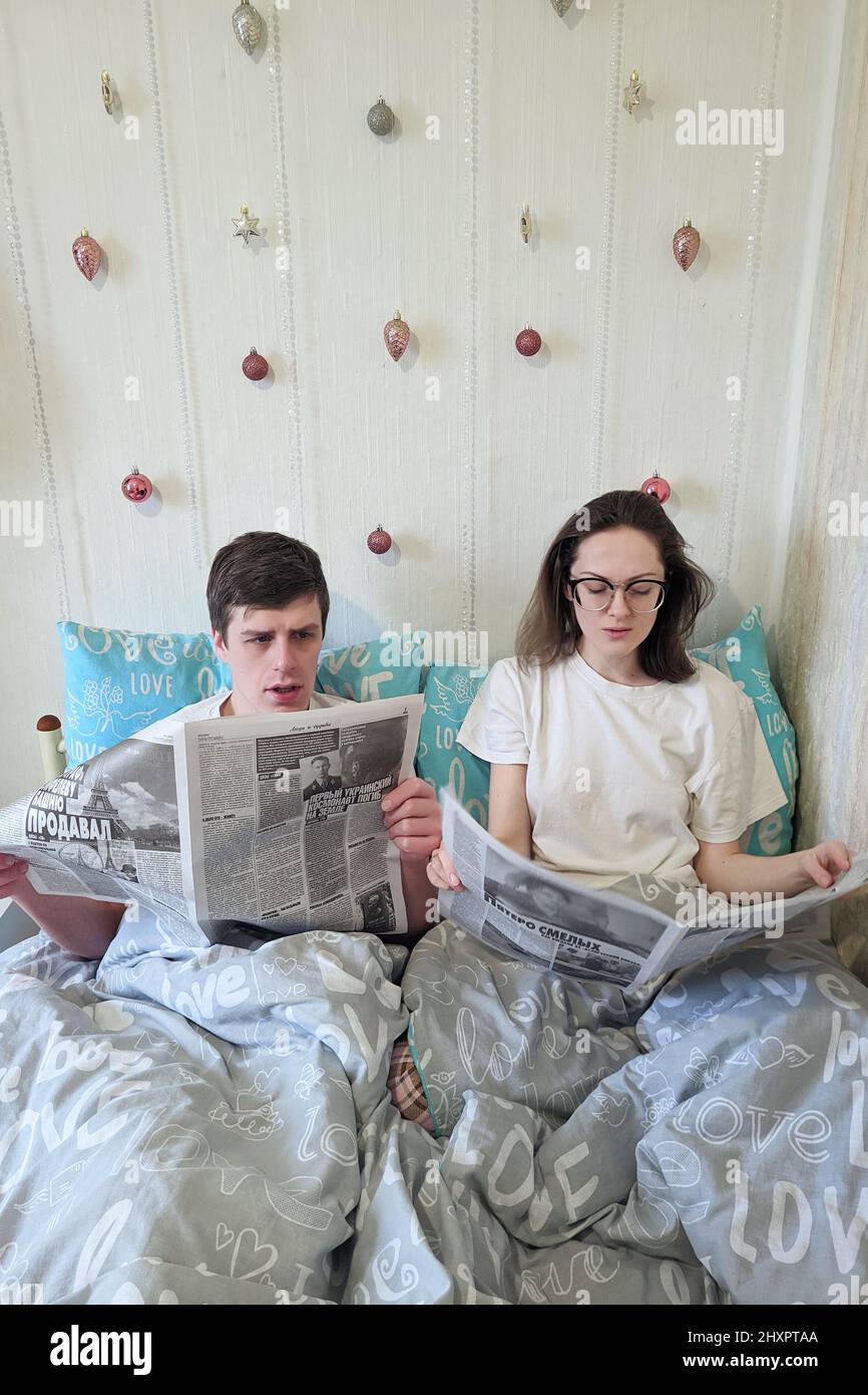 Glückliches Paar, das im Bett in einem gemütlichen Haus liegt, Zeitung liest, Kaffee trinkt. Junger Mann in einem T-Shirt und eine niedliche Frau. Familienurlaub und Zweisamkeit, Stockfoto