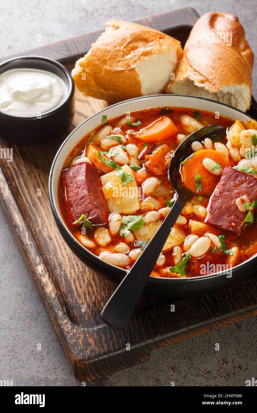 Traditionelle ungarische Babgulyas-Bohnen-Gulaschsuppe aus Pinto-Bohnen, geräuchertem Fleisch, Sellerie, Karotten in der Nähe auf dem Teller auf dem Holztablett. Vertikal Stockfoto