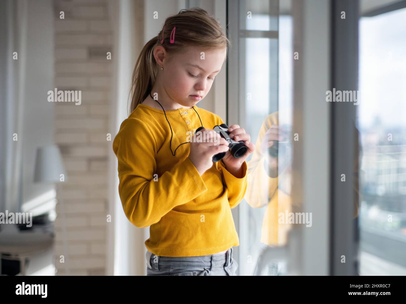 Neugieriges kleines Mädchen mit Down-Syndrom und Fernglas, das zu Hause auf das Fenster schaut. Stockfoto