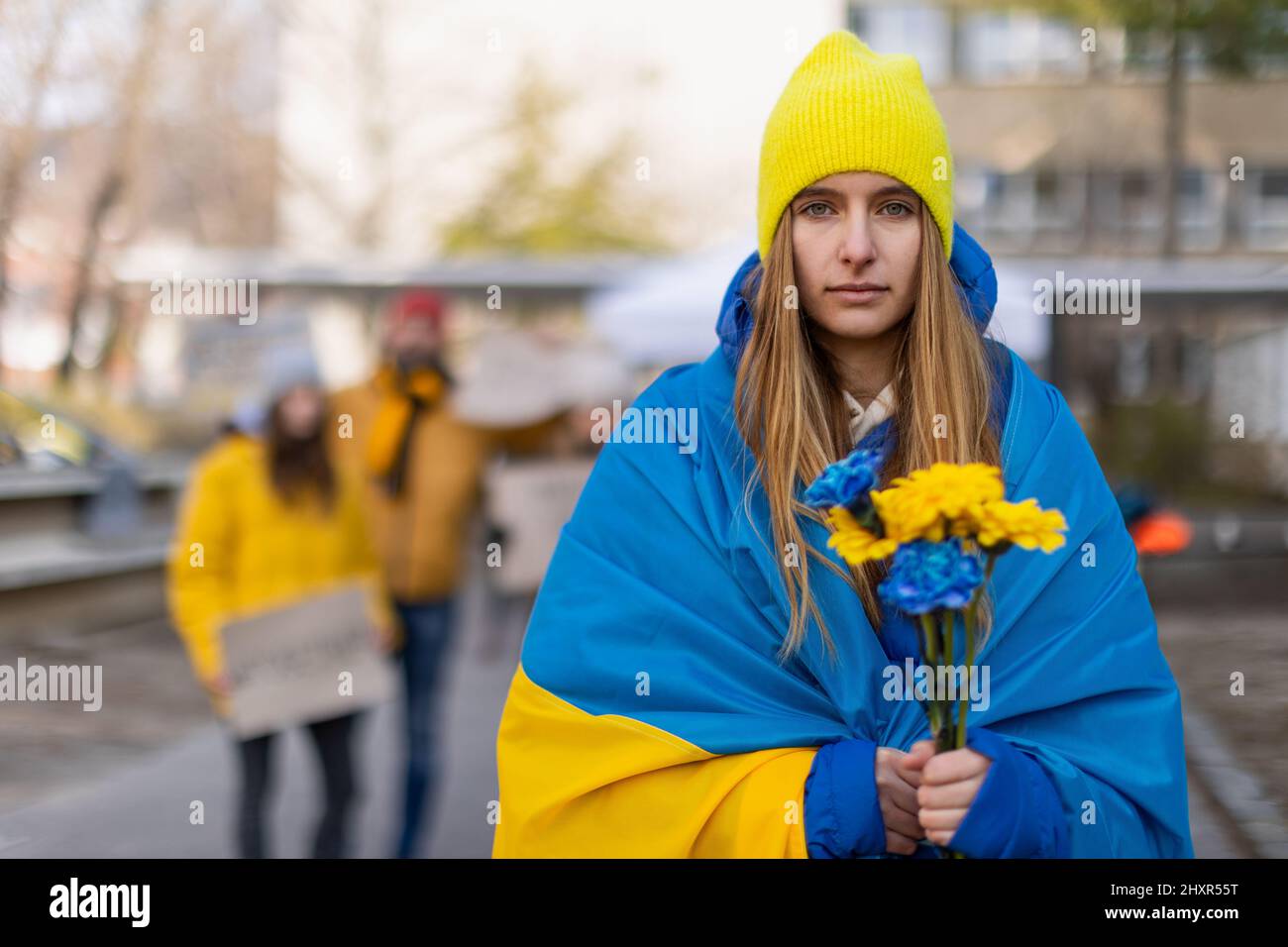 Protest gegen russische Invasion in der Ukraine. Junge Frau in ukrainische Flagge gehüllt mit blauen und gelben Blumen. Stockfoto