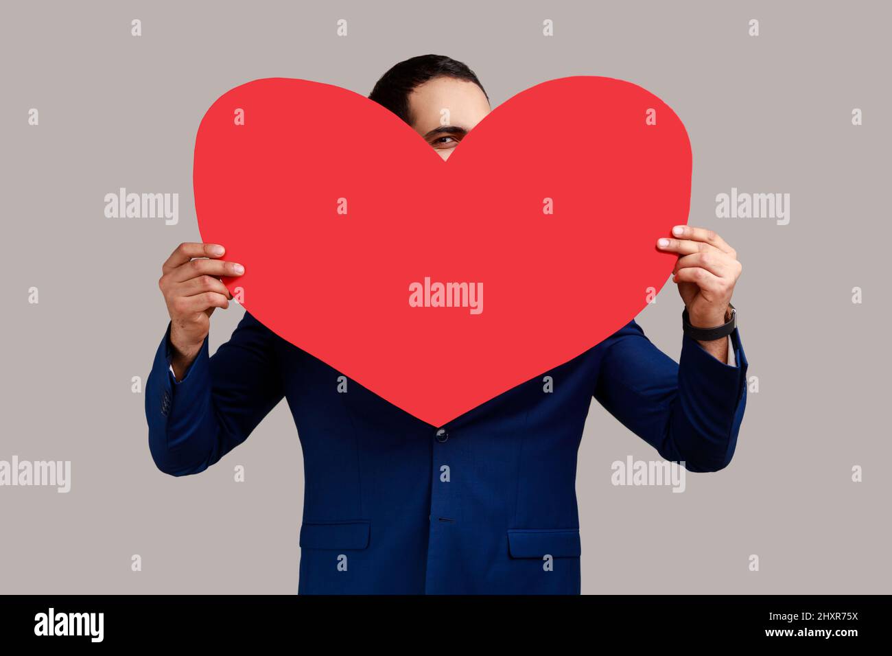Der Mann versteckt sich hinter einem großen roten Herzen und schaut mit neugierigen neugierigen neugierigen Augen auf die Kamera, drückt aus, dass ich dich liebe, Zuneigung und Zuneigung. Innenaufnahme des Studios isoliert auf grauem Hintergrund. Stockfoto