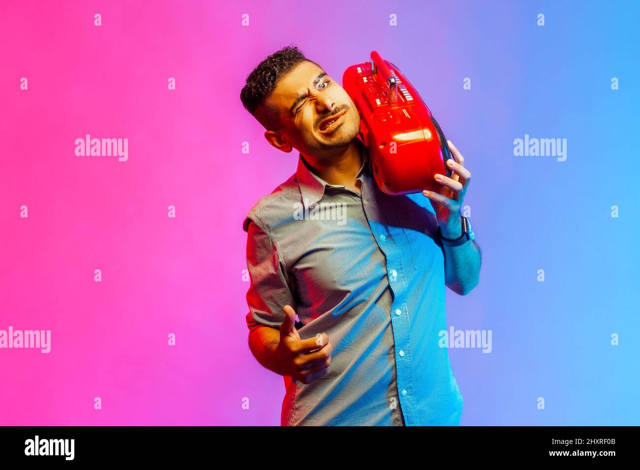 Witziger, hübscher Mann im Hemd, der Musik lauscht, den roten Tonbandrekorder auf der Schulter hält, an der Kamera zwinkert und den Daumen nach oben zeigt. Innenaufnahmen im Studio, isoliert auf farbigem Neonlicht-Hintergrund. Stockfoto