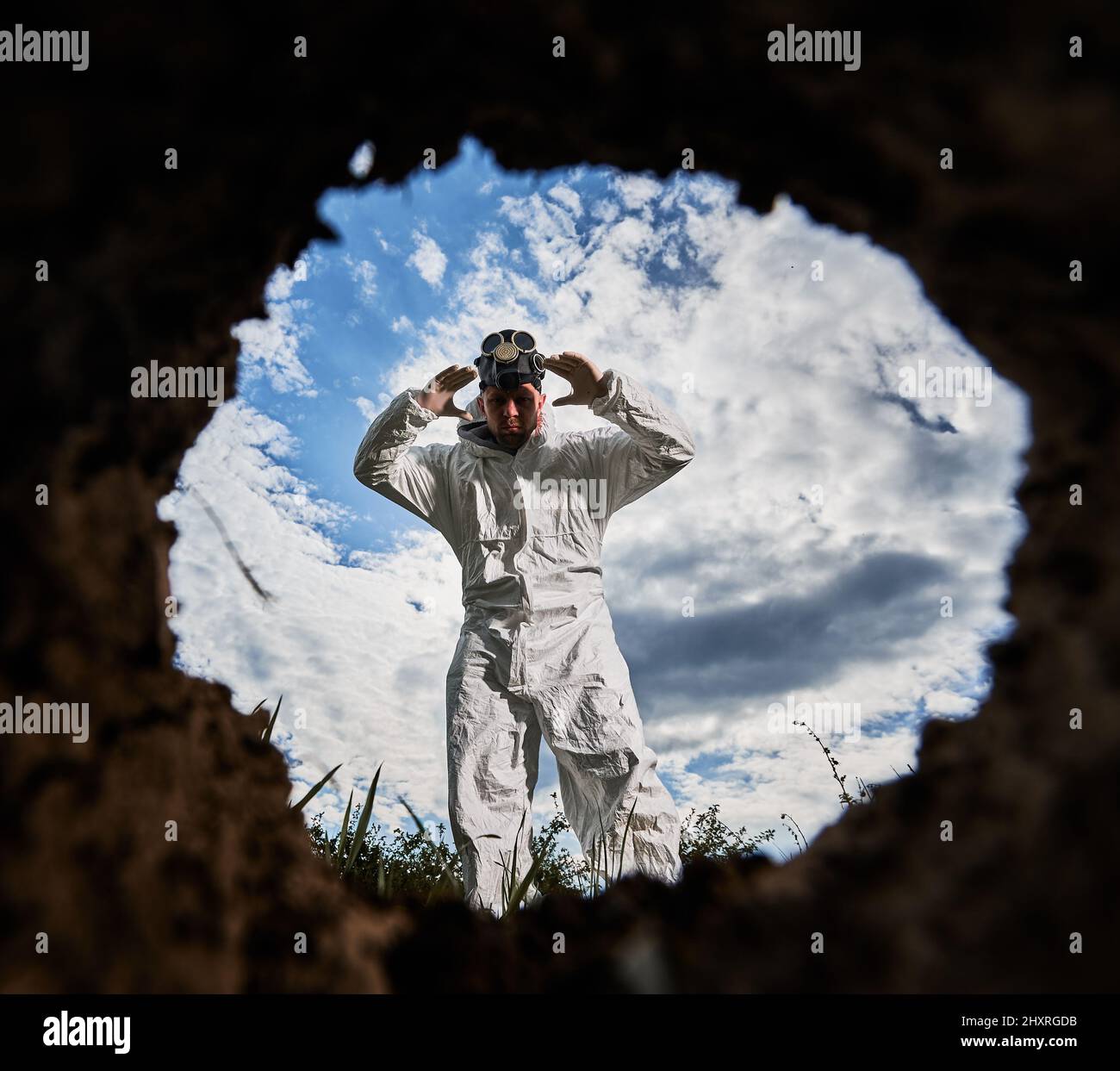 Blick von der Erdgrube des Explorers in weißem Schutzanzug, der gegen den wolkigen Himmel steht, seine Hände zur Gasmaske hochhebt, die er am Kopf trägt und nach unten blickt. Stockfoto