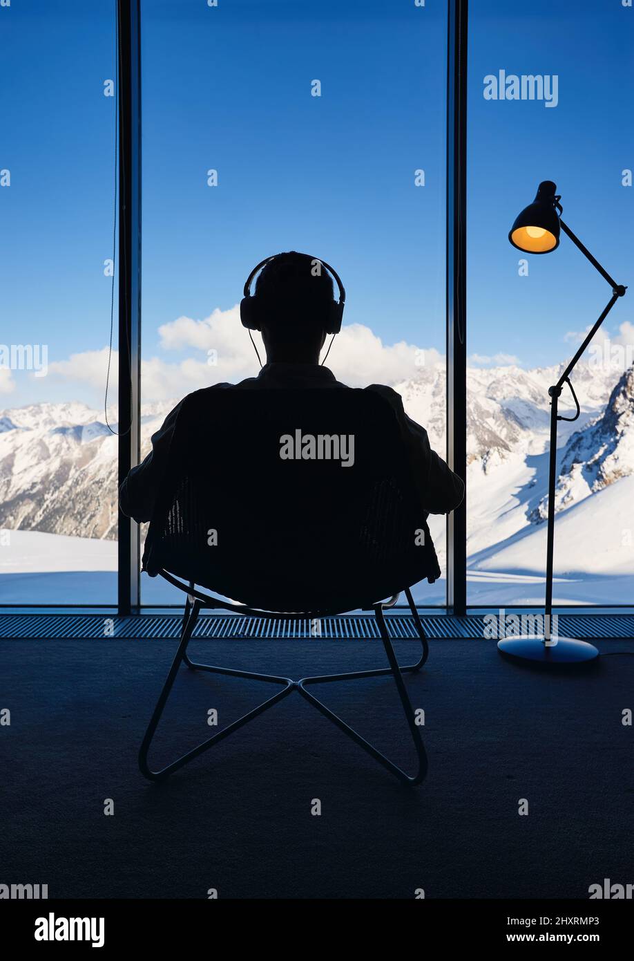 Der Mann in der Silhouette sitzt auf dem Stuhl und hört am Kopfhörer Musik in der Nähe des Panoramafensters mit friedlicher Aussicht auf eine wunderschöne verschneite Berglandschaft. Loft I Stockfoto