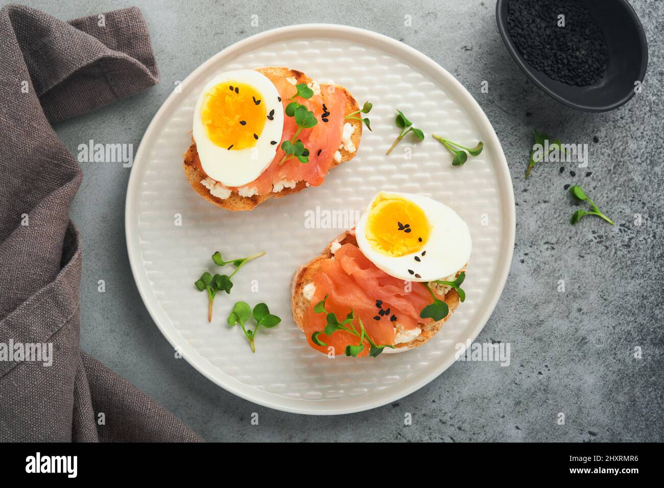 Sandwich mit leckeren Belägen geräucherter Lachs, Eier, Kräuter und Mikrogrüns Rettich, schwarze Sesam-Samen über weißem Teller auf grauem Beton Tisch Backgro Stockfoto