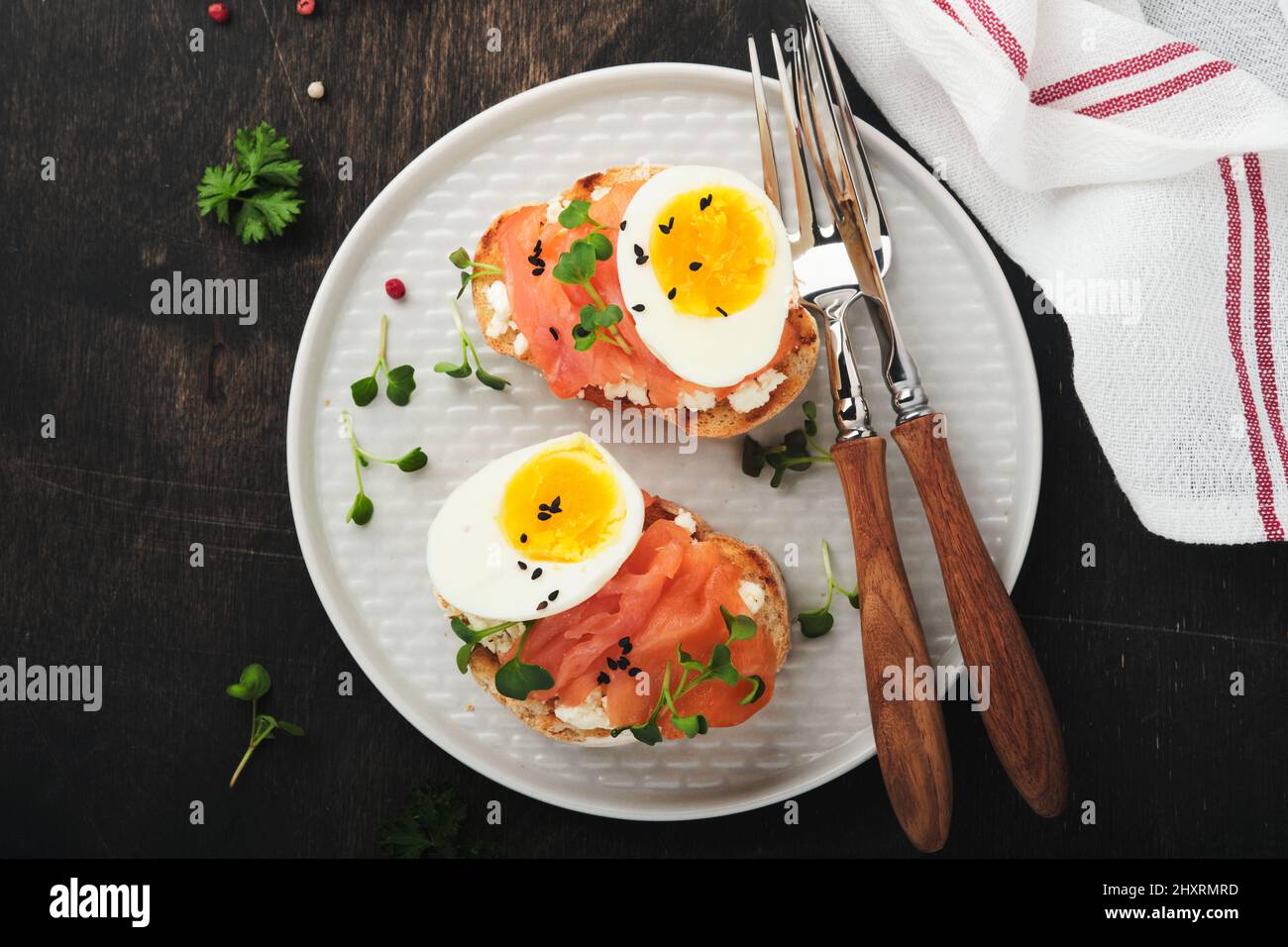 Sandwich mit leckeren Belägen geräucherter Lachs, Eier, Kräuter und Mikrogrüns Rettich, schwarze Sesam-Samen über weißem Teller auf altem Holztischhintergrund Stockfoto