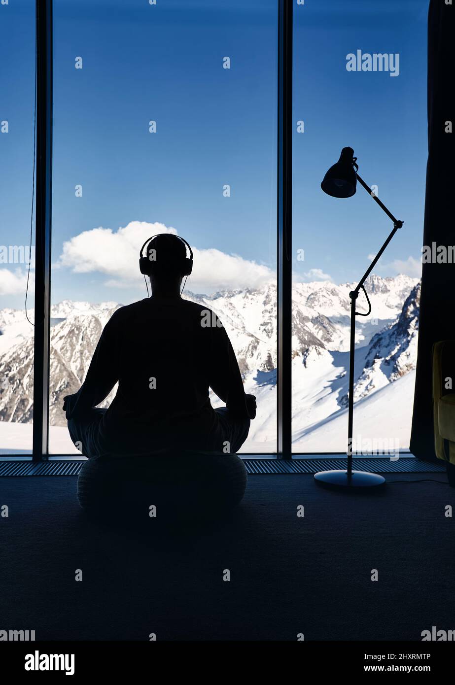 Junge Frau, die Meditationsyogas in Kreuzbeinpose mit Kopfhörern macht, hört Musik im Zimmer mit Panoramablick auf eine wunderschöne Berglandschaft. Hea Stockfoto
