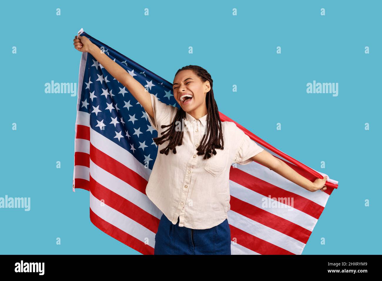 Fröhliche attraktive Frau mit schwarzen Dreadlocks, die die USA-Flagge über den Schultern halten und die Augen geschlossen und fröhlich lächeln, in einem weißen Hemd. Innenaufnahme des Studios isoliert auf blauem Hintergrund. Stockfoto
