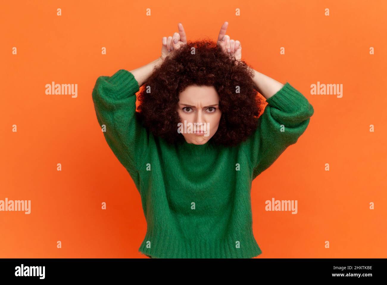 Frau mit Afro-Frisur trägt grünen Pullover sieht bedrohlich und zeigt Bullhorn Zeichen, hält die Finger auf dem Kopf, widerstreitende Person. Innenaufnahme des Studios isoliert auf orangefarbenem Hintergrund. Stockfoto