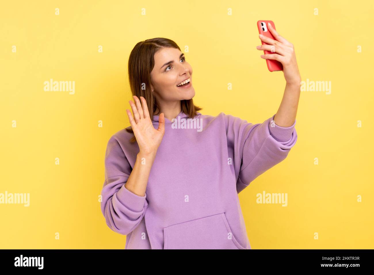 Porträt einer hübschen Frau, die mit der Hand zum Smartphone winkt und Selfie macht, während sie einen Videoanruf abhält und einen violetten Hoodie trägt. Innenaufnahme des Studios isoliert auf gelbem Hintergrund. Stockfoto