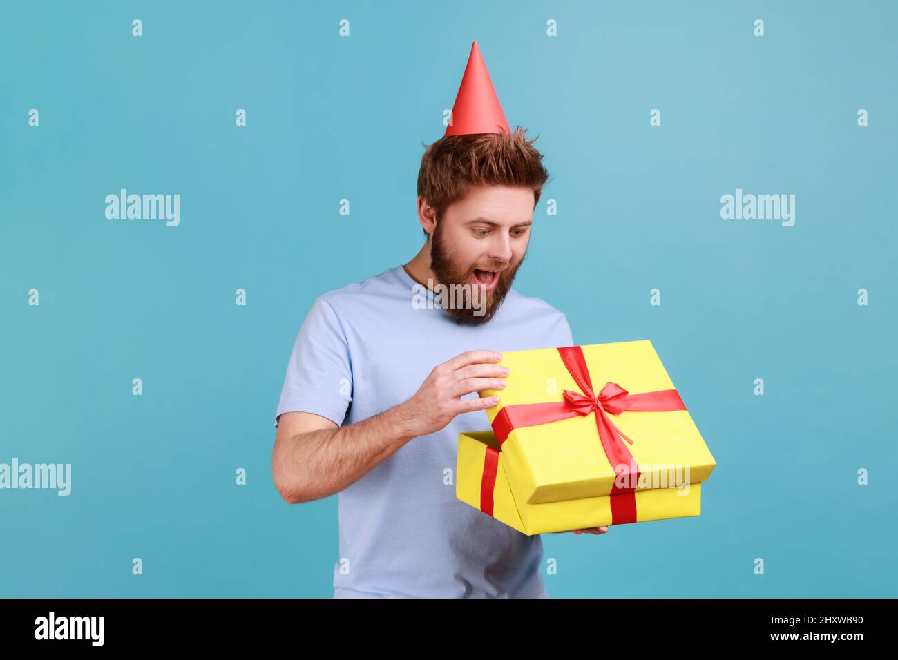 Mann mit Party-Kegel Hut hält geöffnet Geschenkbox und Blick auf die Gegenwart mit aufgeregt aussehen, Auspacken Geschenk, genießen genial Geburtstagsüberraschung. Innenaufnahme des Studios isoliert auf blauem Hintergrund. Stockfoto