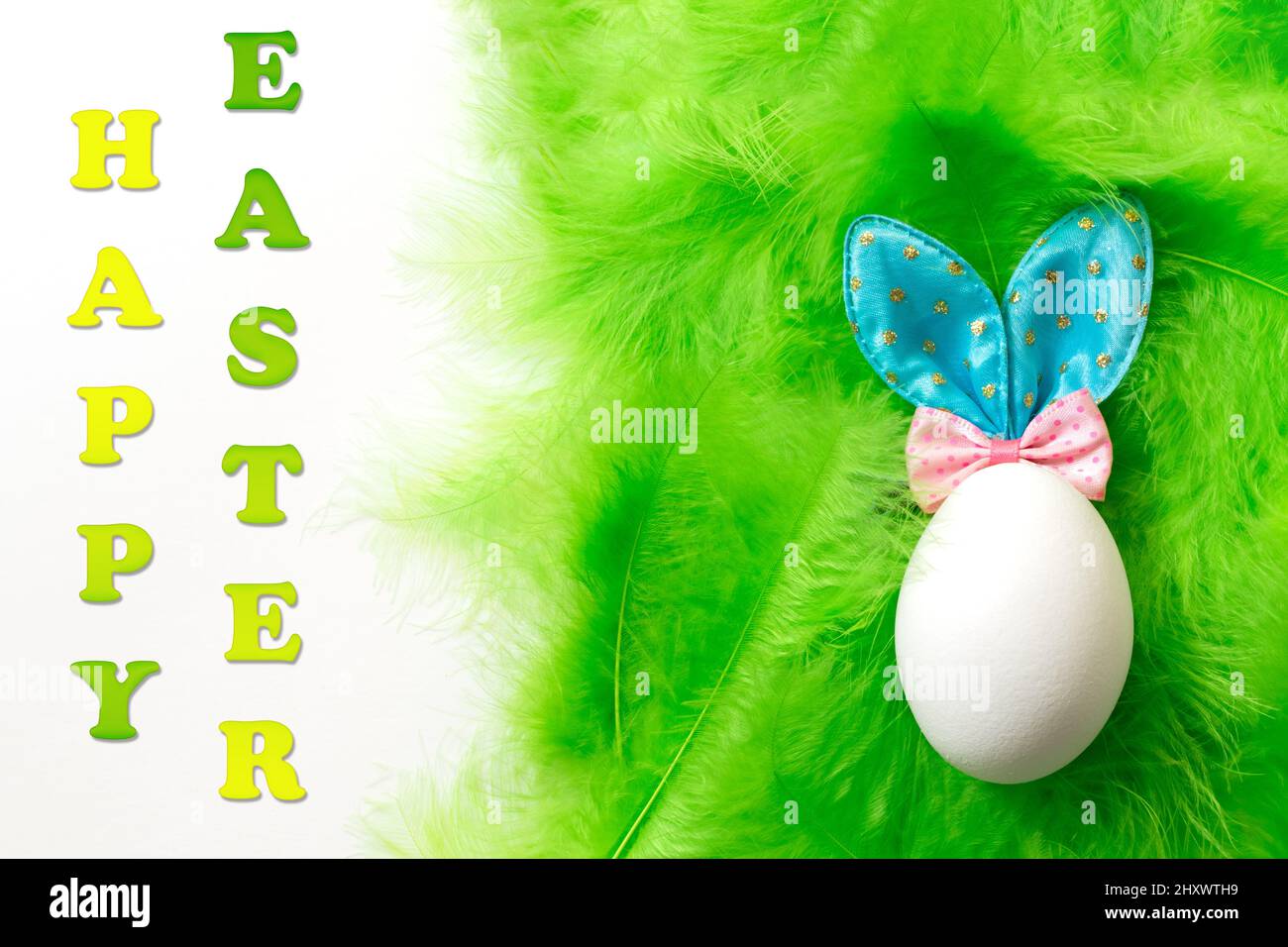 Weißes Ei mit Osterhasen-Ohren auf einem grünen weichen, zarten Federhintergrund und Text Frohe Ostern. Traditionelles Frühlingskonzept. Osterkonzept. Stockfoto
