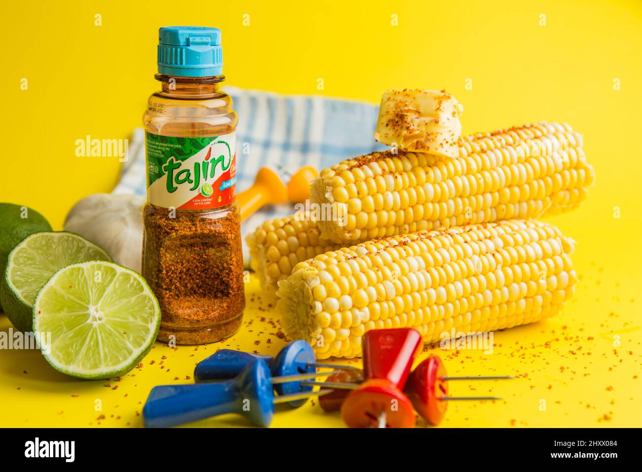 Tajin mexikanische Würze aus Chili, Limette und Meersalz mit frisch geschnittenen Limetten mit Mais auf dem Kohlenmontagesatz, wobei die Spieße auf einem gelben Hintergrund gehalten werden Stockfoto