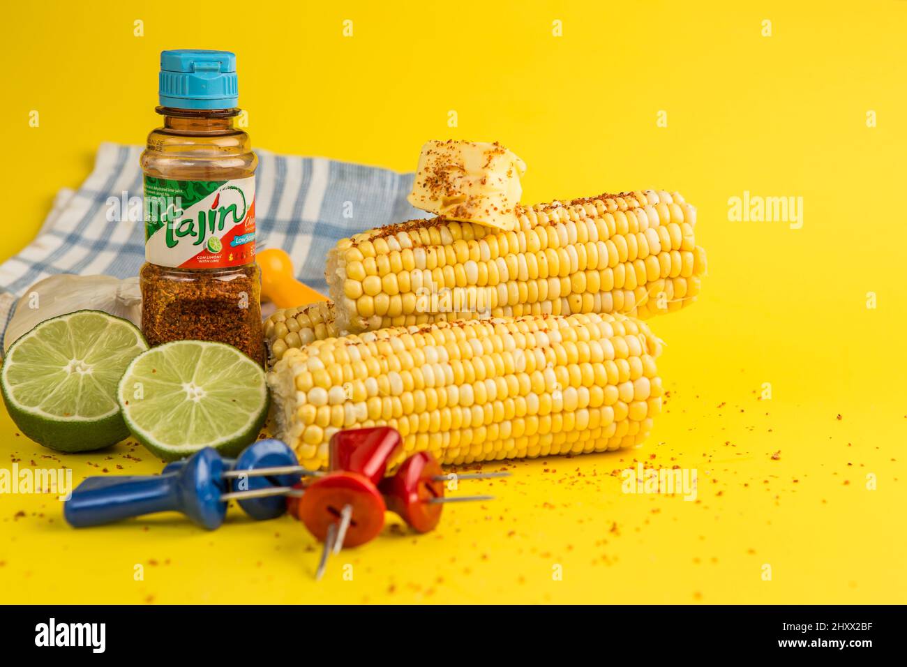 Tajin mexikanische Würze aus Chili, Limette und Meersalz mit frisch geschnittenen Limetten mit Mais auf dem Kohlenmontagesatz, wobei die Spieße auf einem gelben Hintergrund gehalten werden Stockfoto