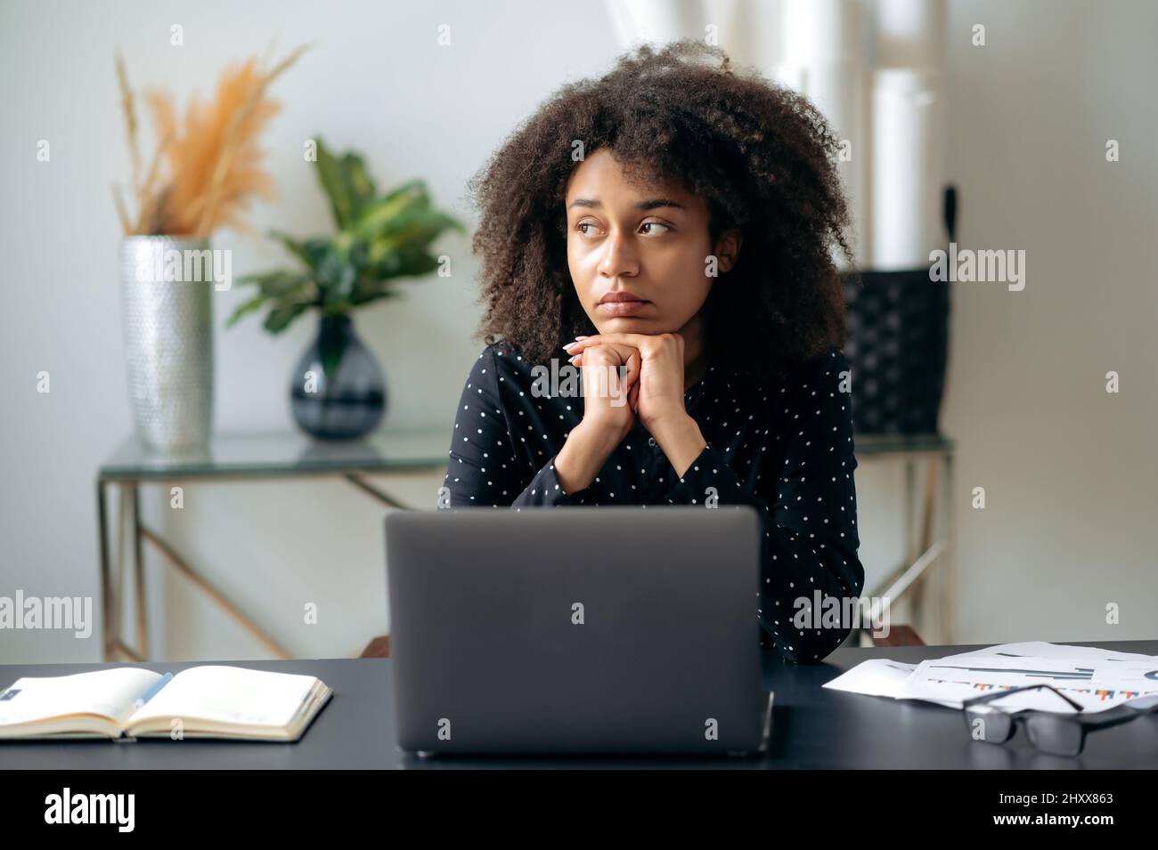 Müde afroamerikanische lockige Frau, Geschäftsfrau, Direktorin, Managerin, sitzt vor einem Laptop an ihrem Schreibtisch, ruht sich von der Computerarbeit aus, schaut zur Seite, träumt von Ruhe oder Urlaub Stockfoto