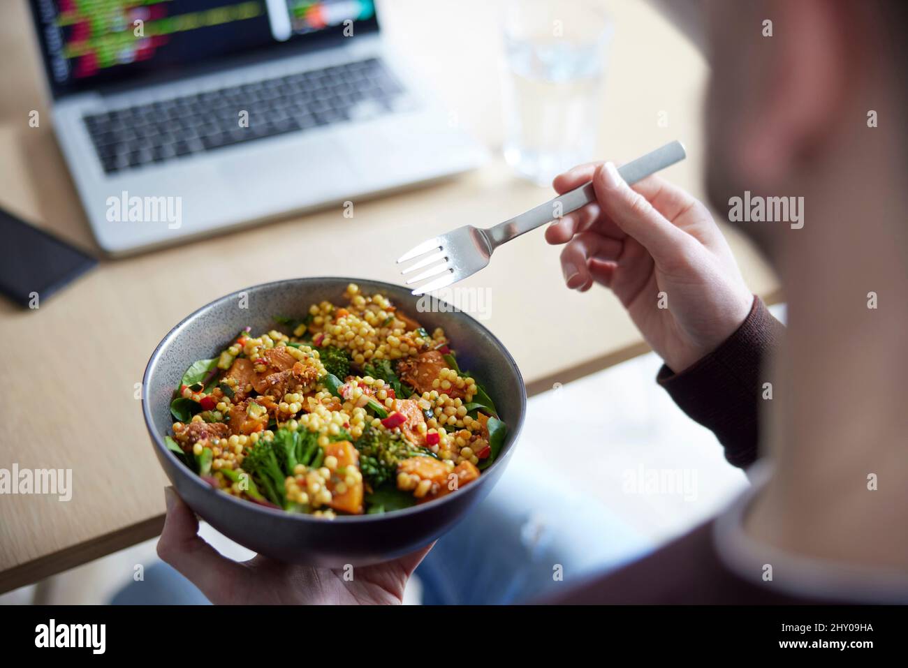 Close Of Man Eating Healthy Vegan Lunch Im Office Deak, Während Man Einen Laptop Benutzt Stockfoto