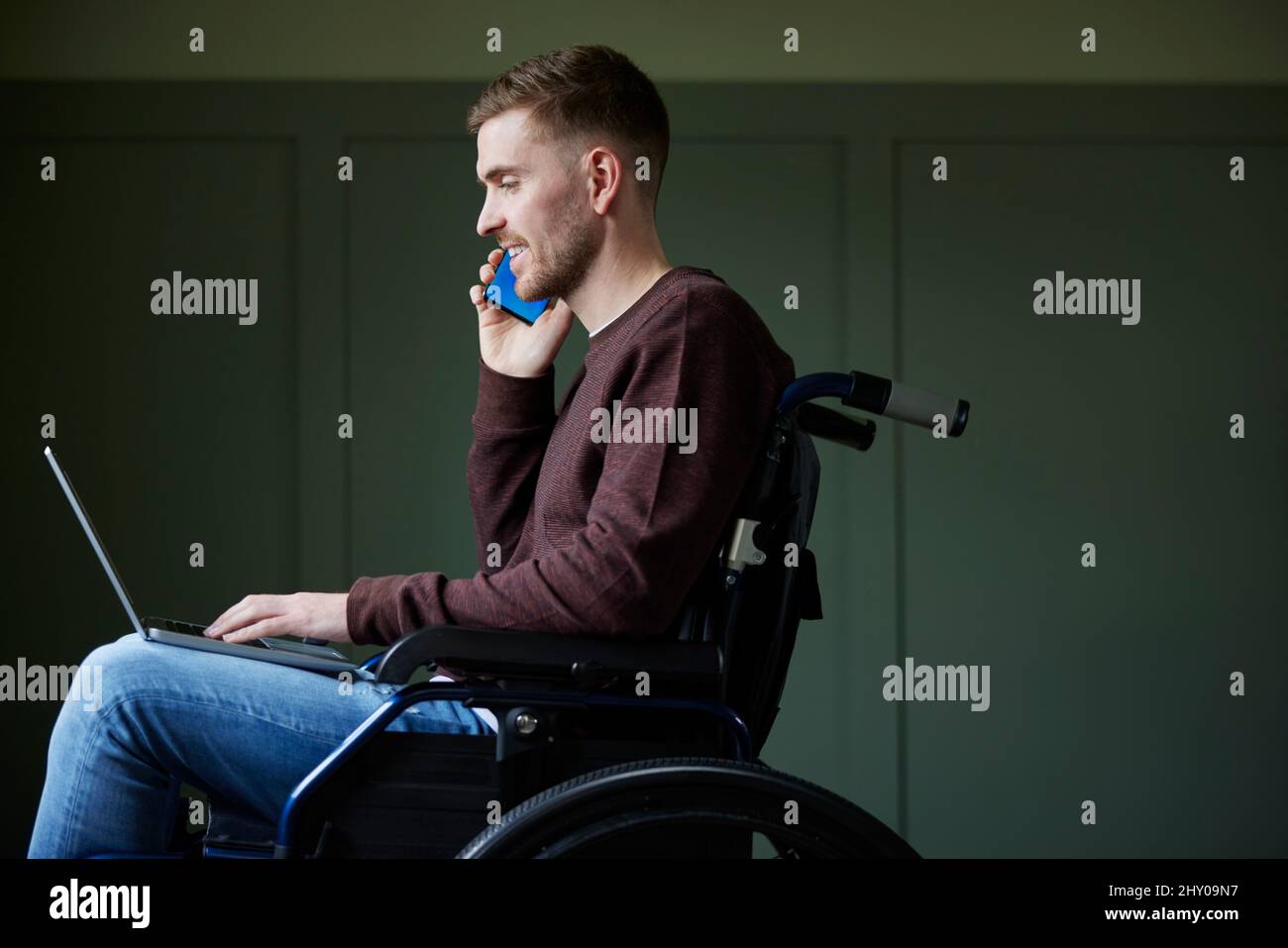 Mann im Rollstuhl, der von zu Hause aus mit Laptop und Mobiltelefon arbeitet Stockfoto