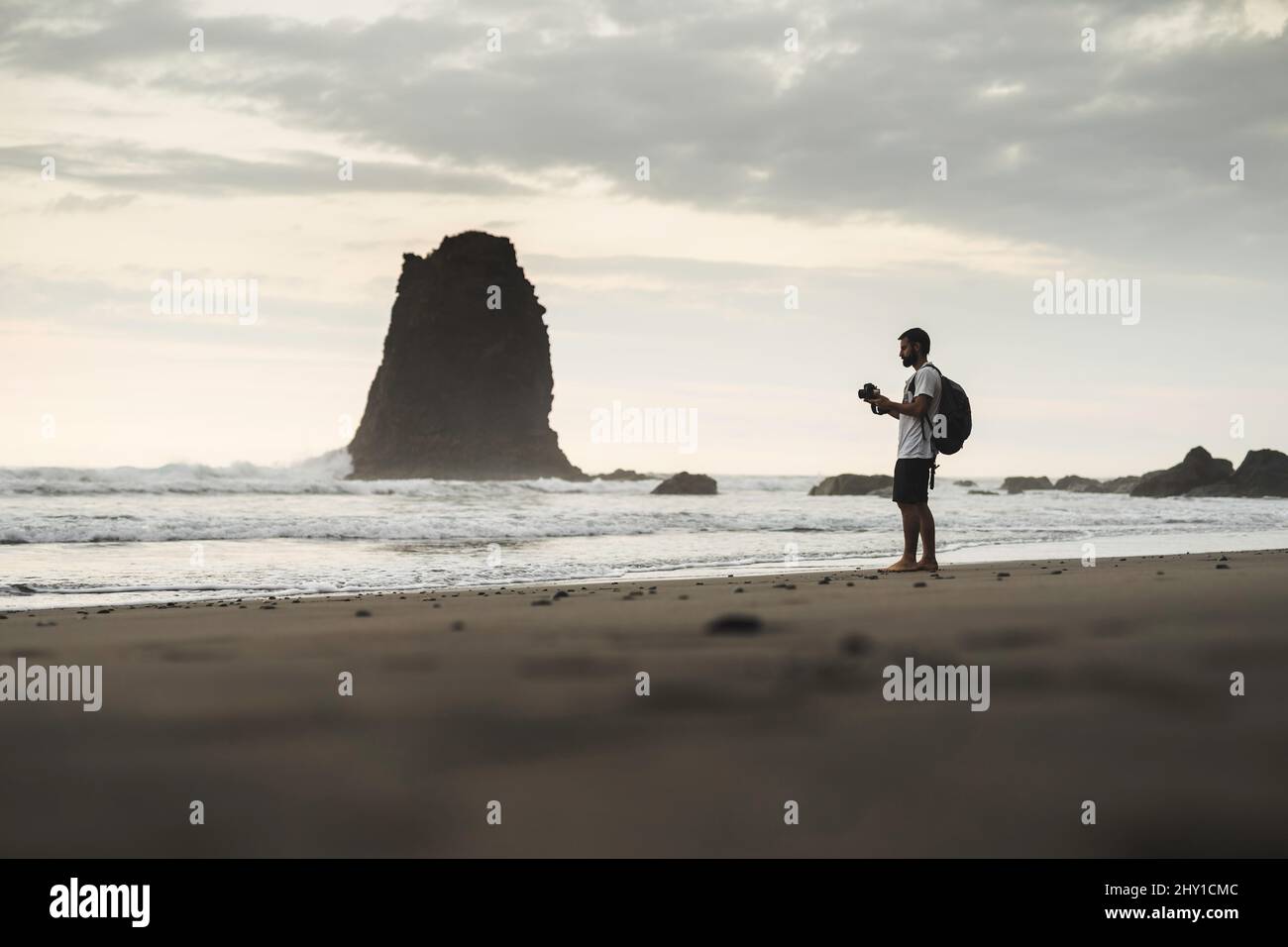 Seitenansicht eines entfernten, nicht erkennbaren männlichen Reisenden, der mit der Fotokamera fotografiert, während er an der Küste in der Nähe des Meeres mit felsiger Formation steht Stockfoto