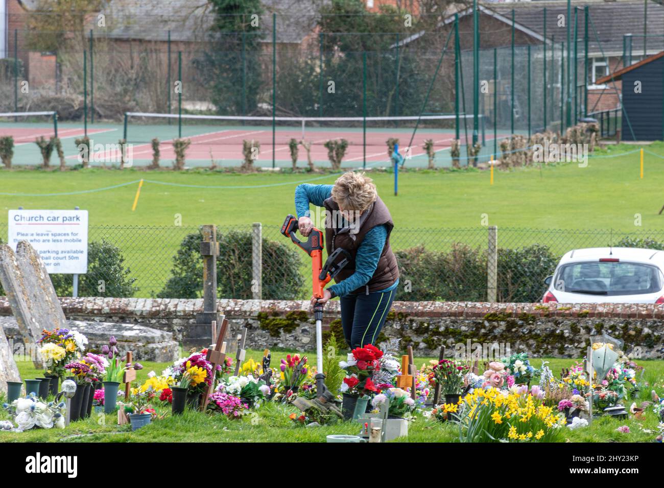 Frau, die in einem Kirchhof langes Gras um Blumen streift und aufräumt, Hampshire, England, Großbritannien Stockfoto