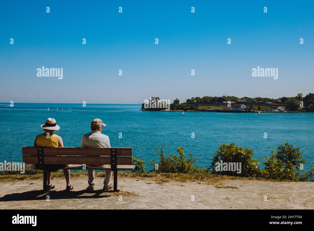 Seniorenpaar sitzt auf einer Holzbank vor dem Meer auf einem blauen Himmel Hintergrund Stockfoto