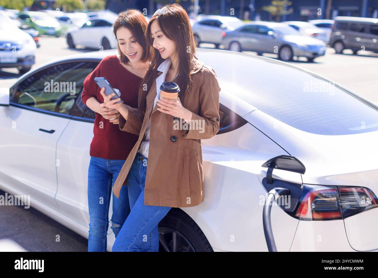 Glückliche junge Frau, die auf dem Stadtparkplatz in der Nähe eines Elektroautos steht, die Autobatterie von einer kleinen Stadtstation lädt, Smartphones beobachtet und nachschaut Stockfoto