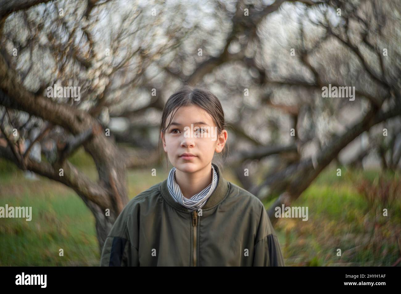 Porträt eines glücklichen Pre-Teen-Mädchens in einem Park. Kind trägt eine grüne Jacke und Rollkragen. Blühende Bäume im Hintergrund. Frühling. Stockfoto