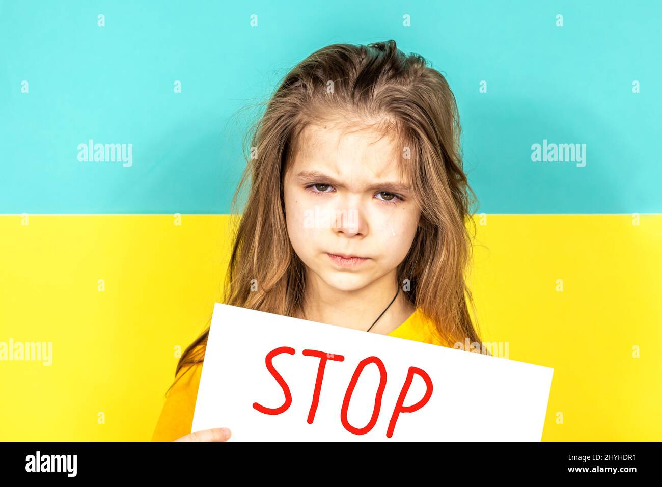 Das Gesicht eines weinenden Mädchens aus der Nähe. Das Mädchen hält vor dem Hintergrund der ukrainischen Flagge ein STOPPPLAKAT in den Händen. Beendet den Krieg in der Ukraine Stockfoto