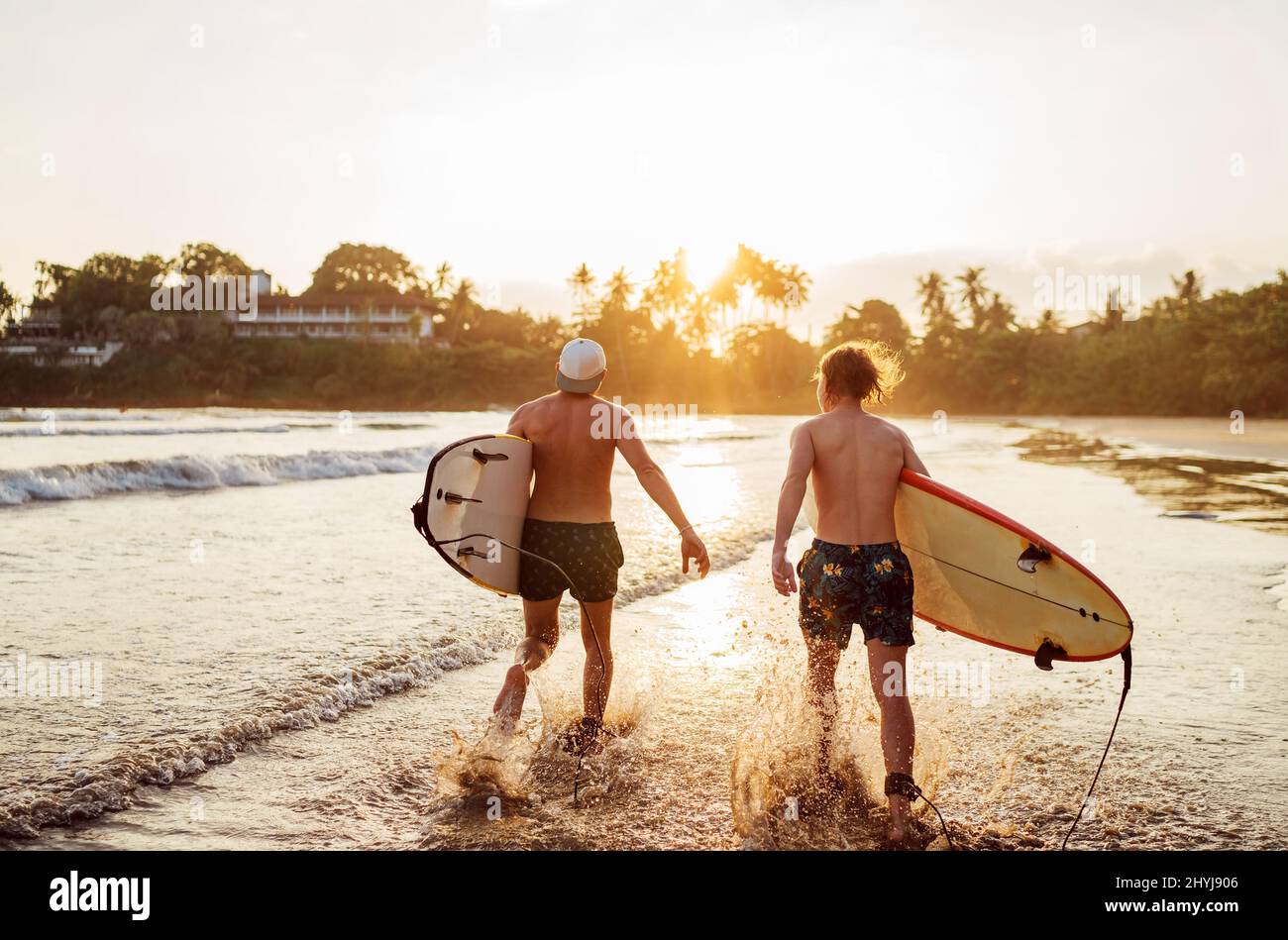 Vater mit Teenager-Sohn, der mit Surfbrettern am Sandstrand mit Palmen im Hintergrund, der von der Sonne des Sonnenuntergangs erhellt wird, entlang läuft. Sie genießen sommerti Stockfoto