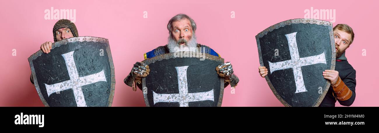 Drei Männer im Mischalter, mittelalterliche Krieger in Rüstung, die sich hinter einem Schild verbergen, der über einem rosafarbenen Studiohintergrund isoliert ist. Schaut erschrocken aus. Collage Stockfoto