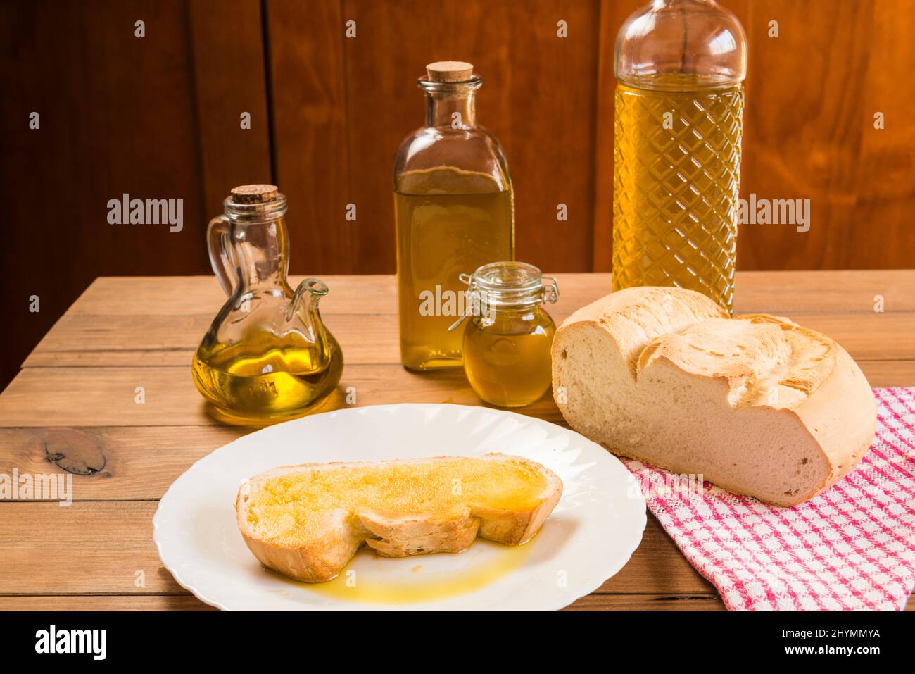 Stillleben einer Scheibe Brot mit nativem Olivenöl extra auf einem weißen Teller, neben Flaschen und Behältern mit Öl gefüllt, auf einem rustikalen Holztisch. Stockfoto
