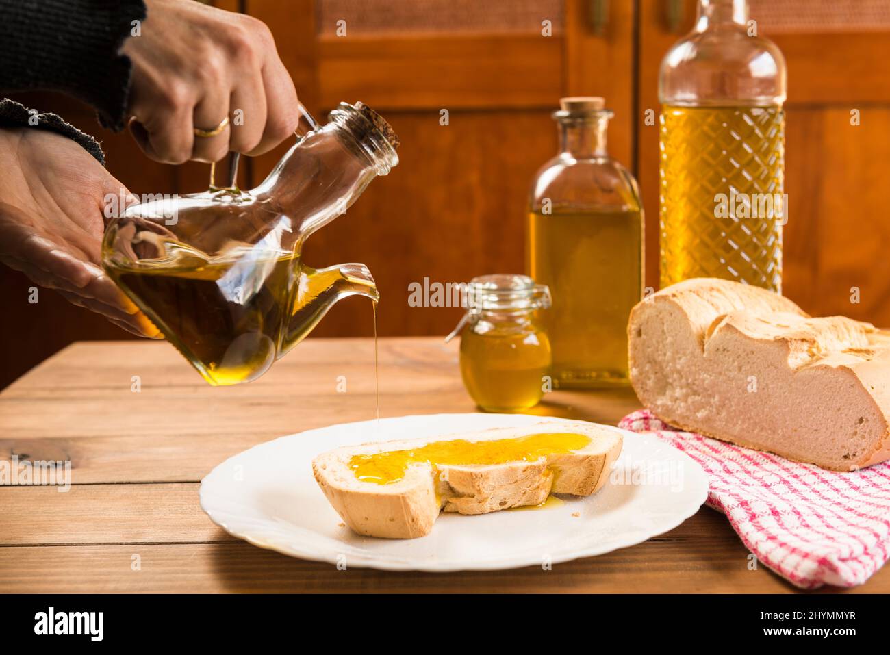 Detail der Hände, die natives Olivenöl extra über eine Scheibe Brot auf einem weißen Teller auf einem rustikalen Holztisch gießen. Stockfoto