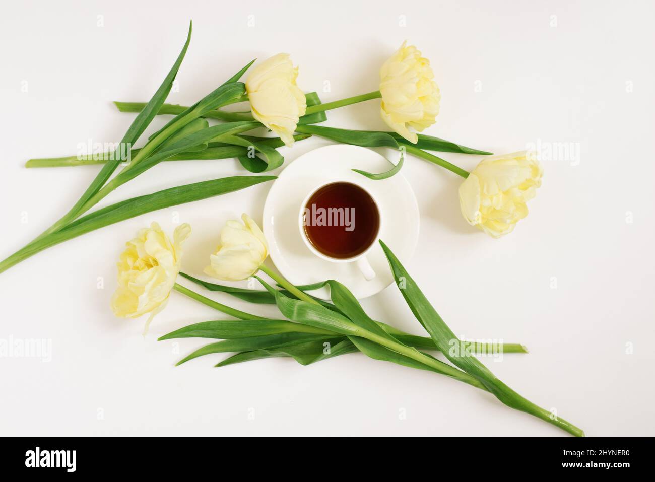 Ein Bouquet aus zarten gelben Tulpen und einer Tasse Tee auf weißem Hintergrund. Draufsicht. Das Konzept eines Blogs oder Freiberuflers. Osterkarte Stockfoto