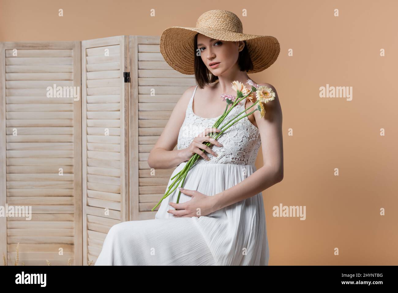 Schwanger Frau in Kleid hält Blumen in der Nähe von Faltschiebe isoliert auf beige Stockfoto